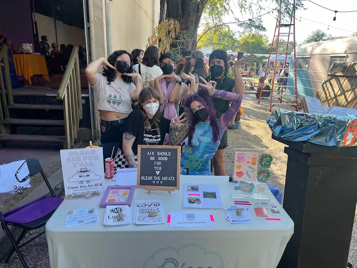 A group of five people gathered together behind a table outside. All five are wearing KN95 or N95 masks. The table has Clear the Air merch on it, along with flyers.