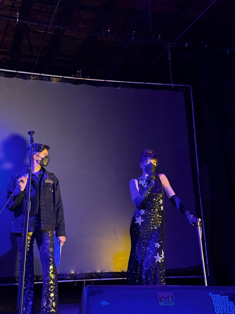 Two people in celestially-themed outfits standing on stage wearing KN95 masks and holding microphones. One of the presenters is leaning on her pink cane.